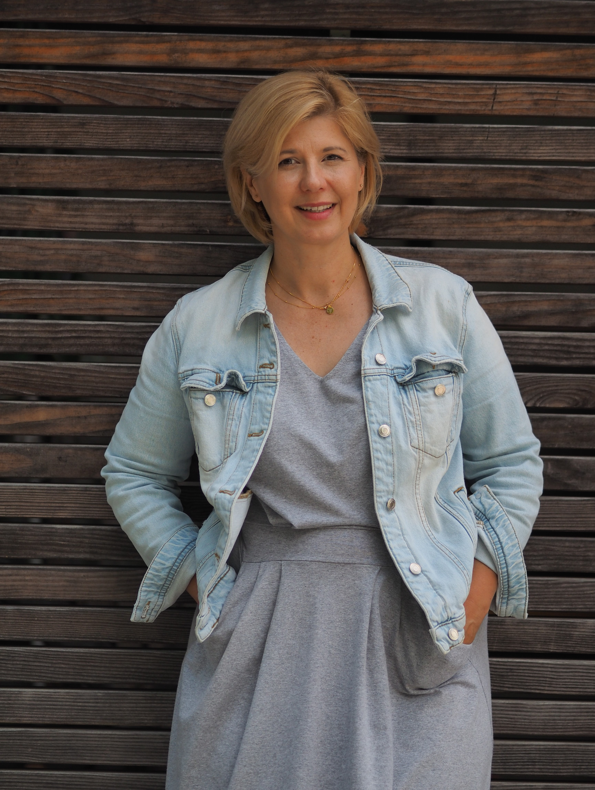 Kobieta o jasnych włosach, w jasnej jeansowej kurtce i szarej sukience, uśmiecha się, stojąc na tle drewnianej ściany.