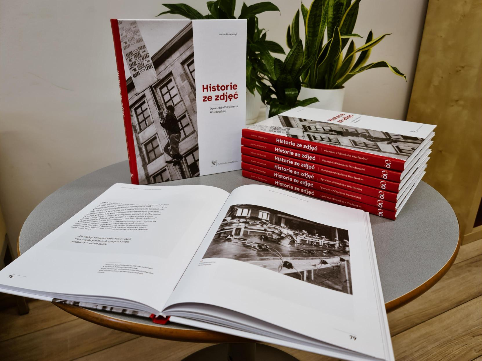 Stos książek "Historie ze zdjęć" na szarym stoliku, obok otwarta strona z historyczną, czarno-białą fotografią oraz doniczkowa roślina.