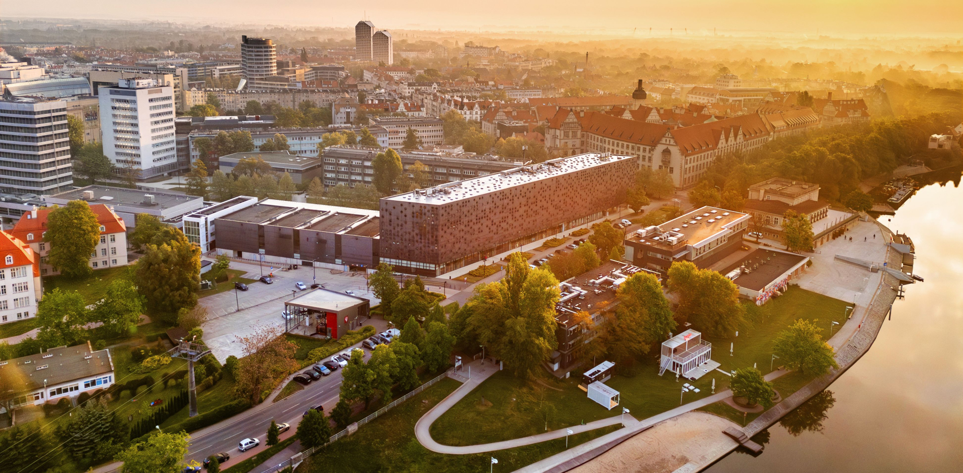 Widok z lotu ptaka na kampus Politechniki Wrocławskiej o wschodzie słońca. Obok nowoczesnych budynków widać zabytkowe gmachy, drzewa i rzekę Odrę. W tle rozciąga się panorama miasta.
