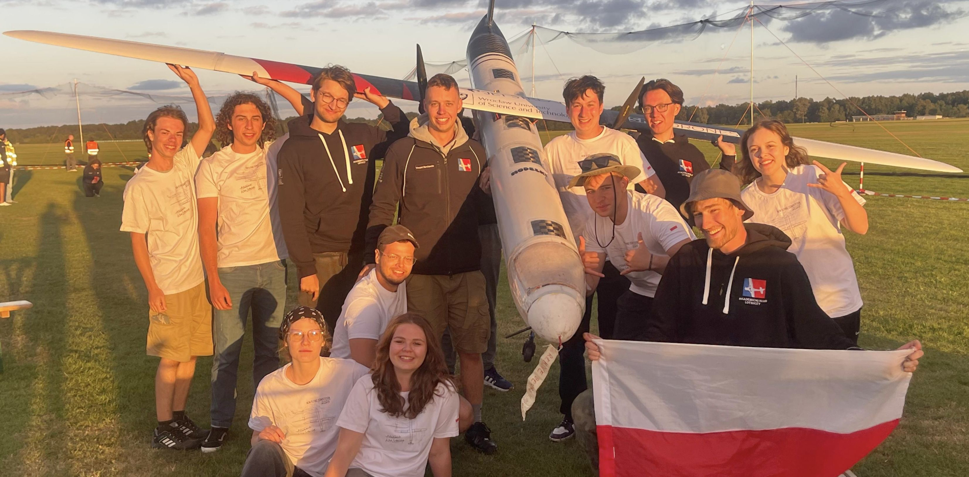 Grupowe zdjęcie studentów z zespołu AKL, stojących na lotnisku. W tle widoczny jest model samolotu z dużymi skrzydłami oraz zachód słońca. Studenci trzymają polską flagę.