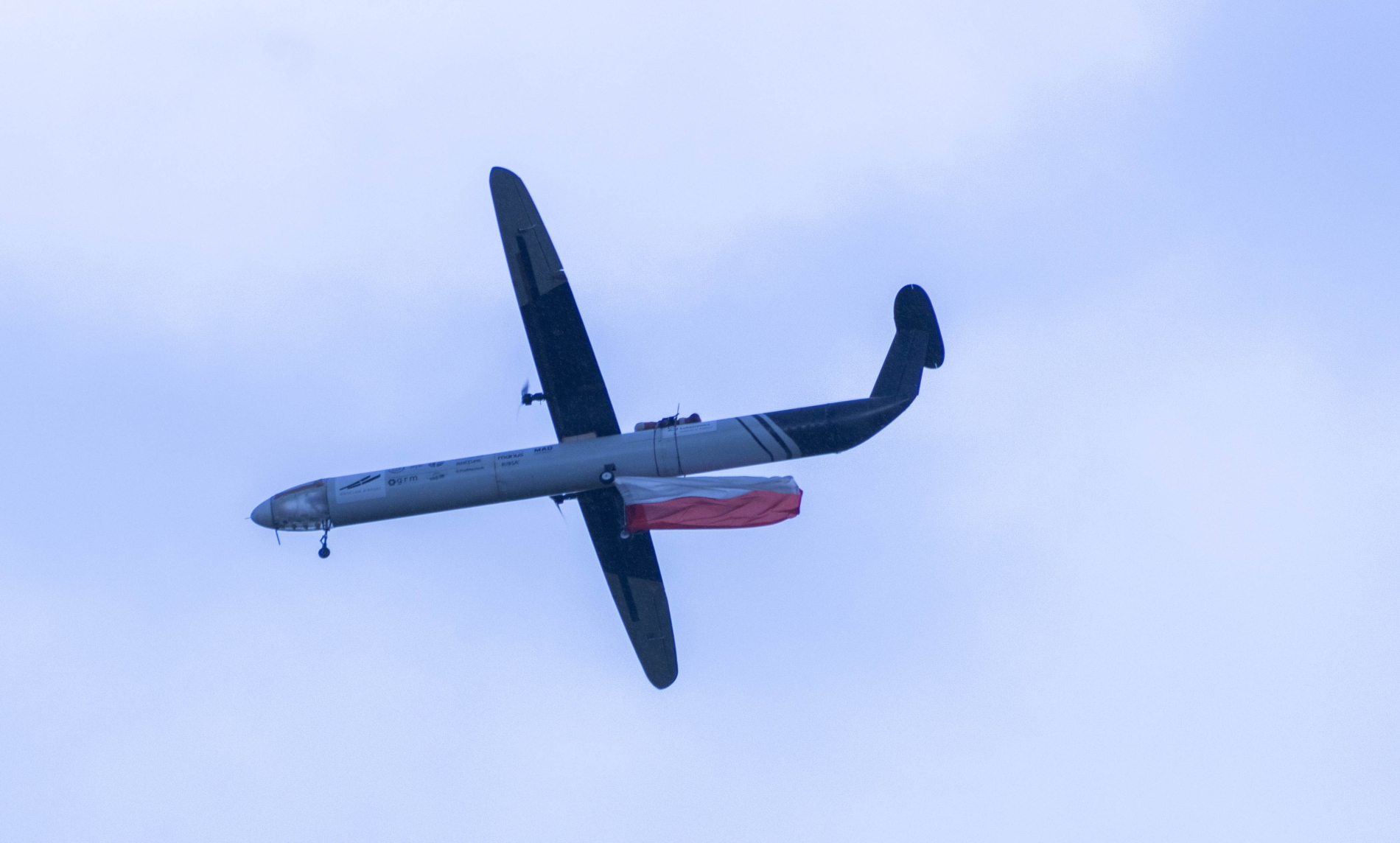 Widok od spodu na szary, bezzałogowy statek powietrzny (dron), który unosi się na tle błękitnego nieba. Pod kadłubem drona zwisa biało-czerwona flaga.