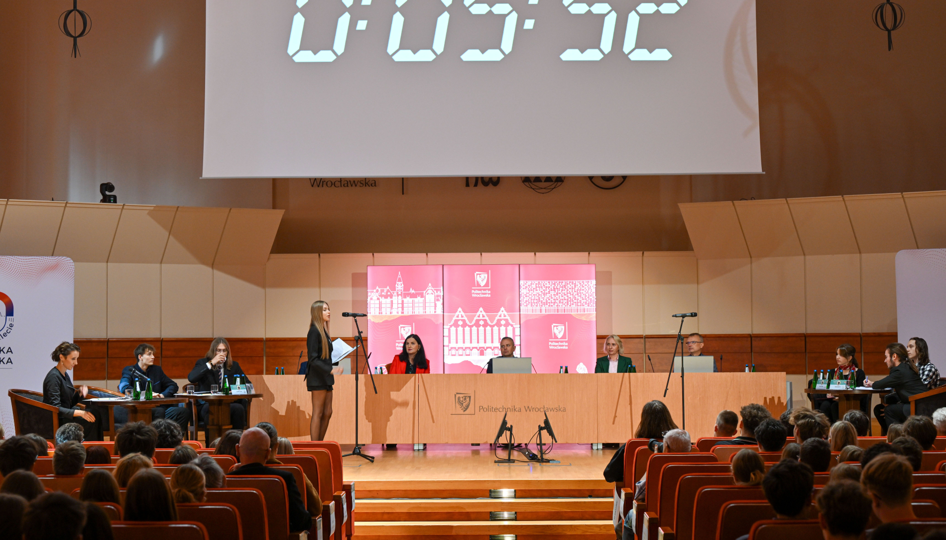 Duża grupa ludzi siedzi na krzesłach w sali audytoryjnej. Na podwyższeniu stoi duży, drewniany stół z logiem Politechniki Wrocławskiej, za którym zasiada panel złożony z dziewięciu osób. Jedna z kobiet stoi przy mównicy. Nad sceną widoczny jest ekran z cyfrowym zegarem.