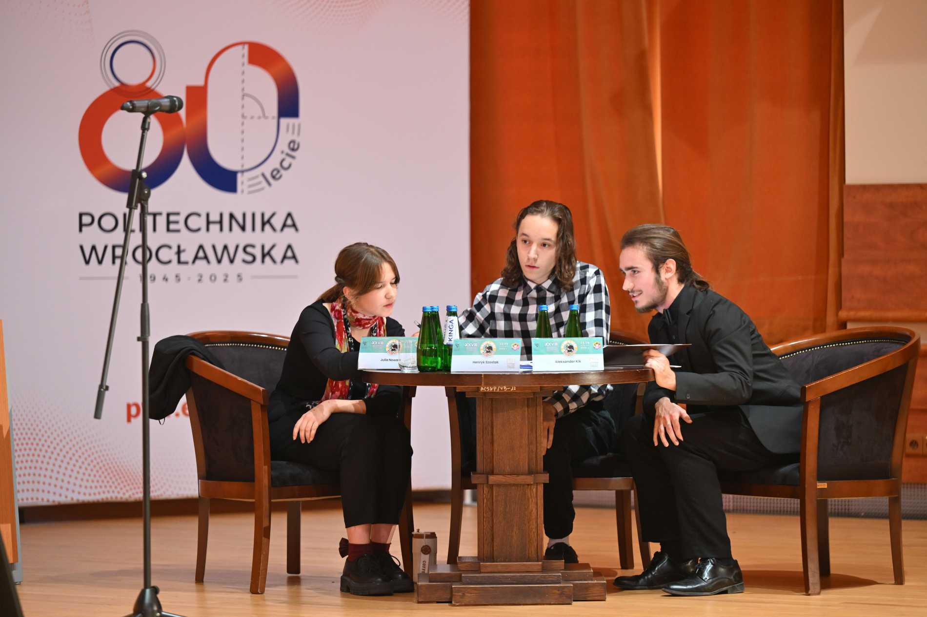 Trzy młode osoby siedzą przy okrągłym, drewnianym stole na podwyższeniu. Po lewej stronie widać fragment mikrofonu na statywie. Za nimi znajduje się plansza z logo Politechniki Wrocławskiej i napisem "80-lecie Politechnika Wrocławska 1945-2025".