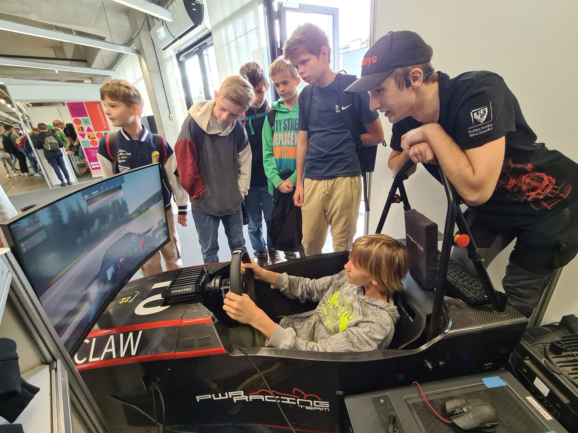 Grupa dzieci i nastolatków stoi wokół symulatora wyścigowego bolidu na stoisku PWR Racing Team. Jedno z dzieci siedzi za kierownicą, a starszy chłopak w czapce pochyla się i pomaga mu. W tle widać innych uczestników wydarzenia.