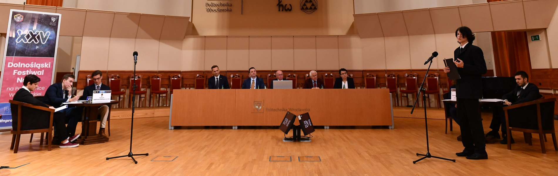Panorama dużej sali wykładowej. Przy długim, drewnianym stole siedzi jury, a z boku, przy mniejszych stolikach, uczestnicy debaty. Młody mężczyzna w czarnym garniturze stoi na środku, trzymając w ręku notatki i mówiąc do mikrofonu.