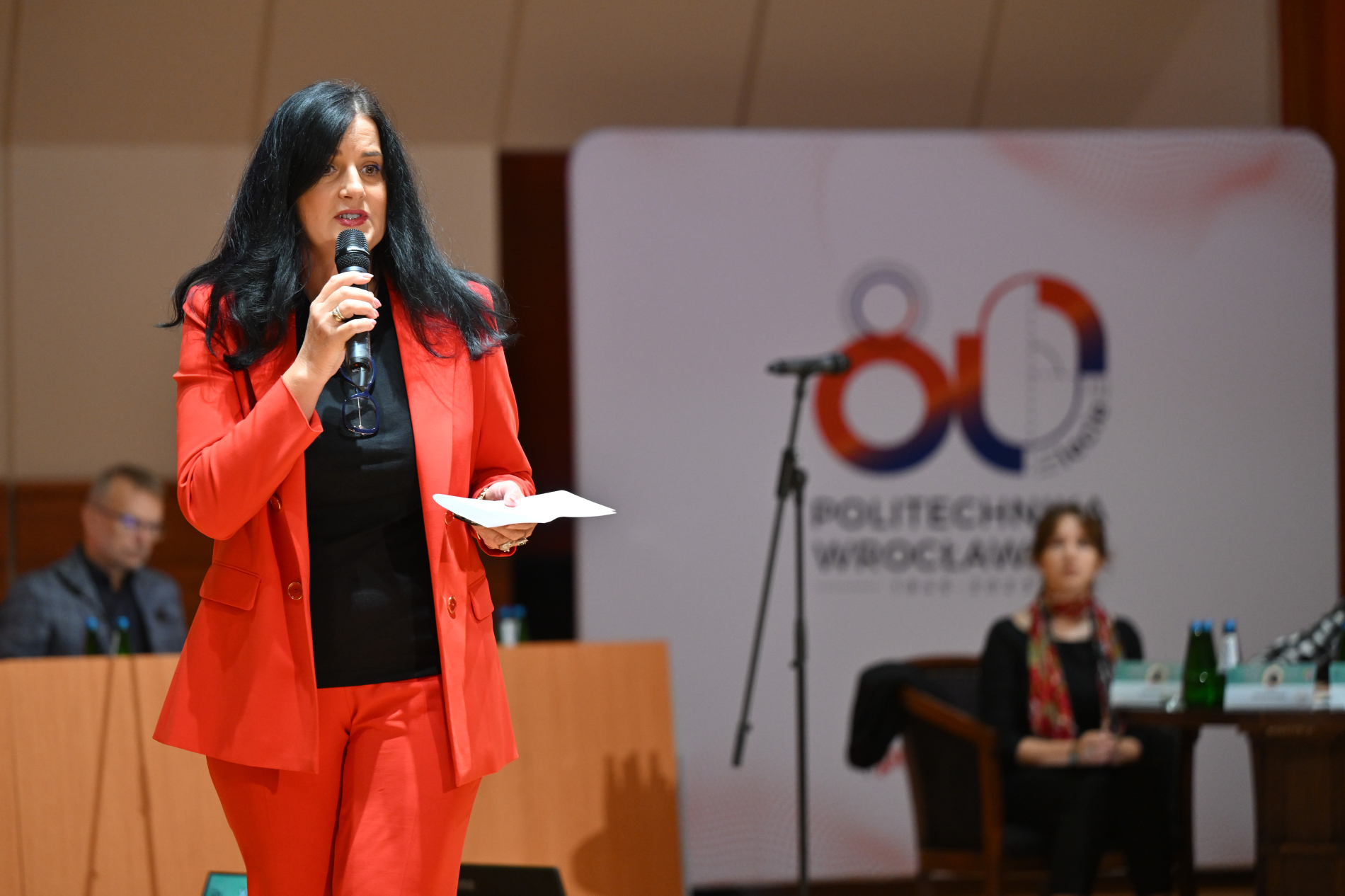 Kobieta w pomarańczowym garniturze, z długimi, czarnymi włosami, stoi przed mikrofonem i trzyma w rękach kartkę. W tle znajduje się baner z logo Politechniki Wrocławskiej i napisem "80-lecie". Po prawej stronie widać fragment siedzącej kobiety, a w lewym dolnym rogu plecy siedzącego mężczyzny.
