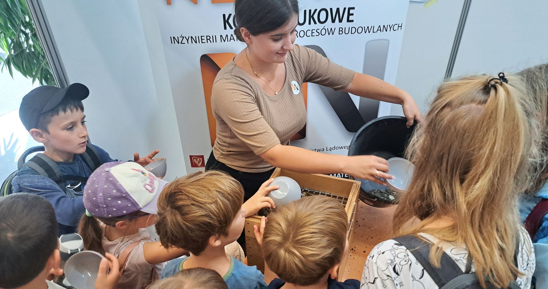 Młoda kobieta w beżowej bluzce z kucykiem, pochyla się nad drewnianą skrzynką, pokazując coś grupie dzieci. Dzieci trzymają miseczki, wpatrując się z zaciekawieniem w to, co znajduje się w skrzynce.
