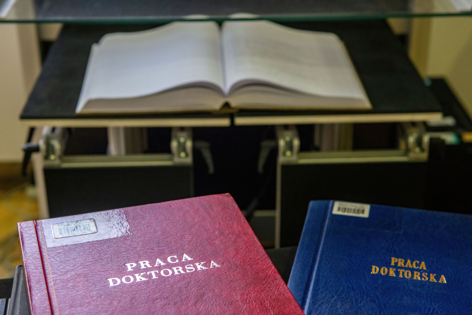 Dwie oprawione prace doktorskie, czerwona i niebieska, leżące na biurku. W tle na podwyższeniu otwarta książka.