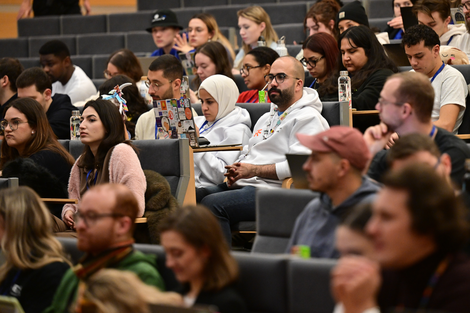 Zbliżenie na dużą grupę młodych ludzi, uczestników konferencji, siedzących w rzędach. Skupiają się oni na prelekcji