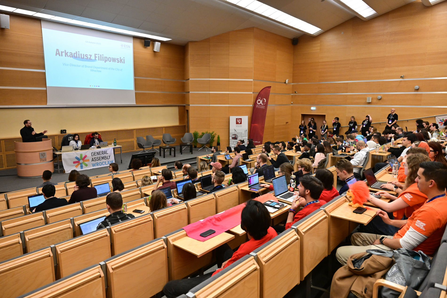 Sala konferencyjna wypełniona uczestnikami siedzącymi w rzędach z laptopami.