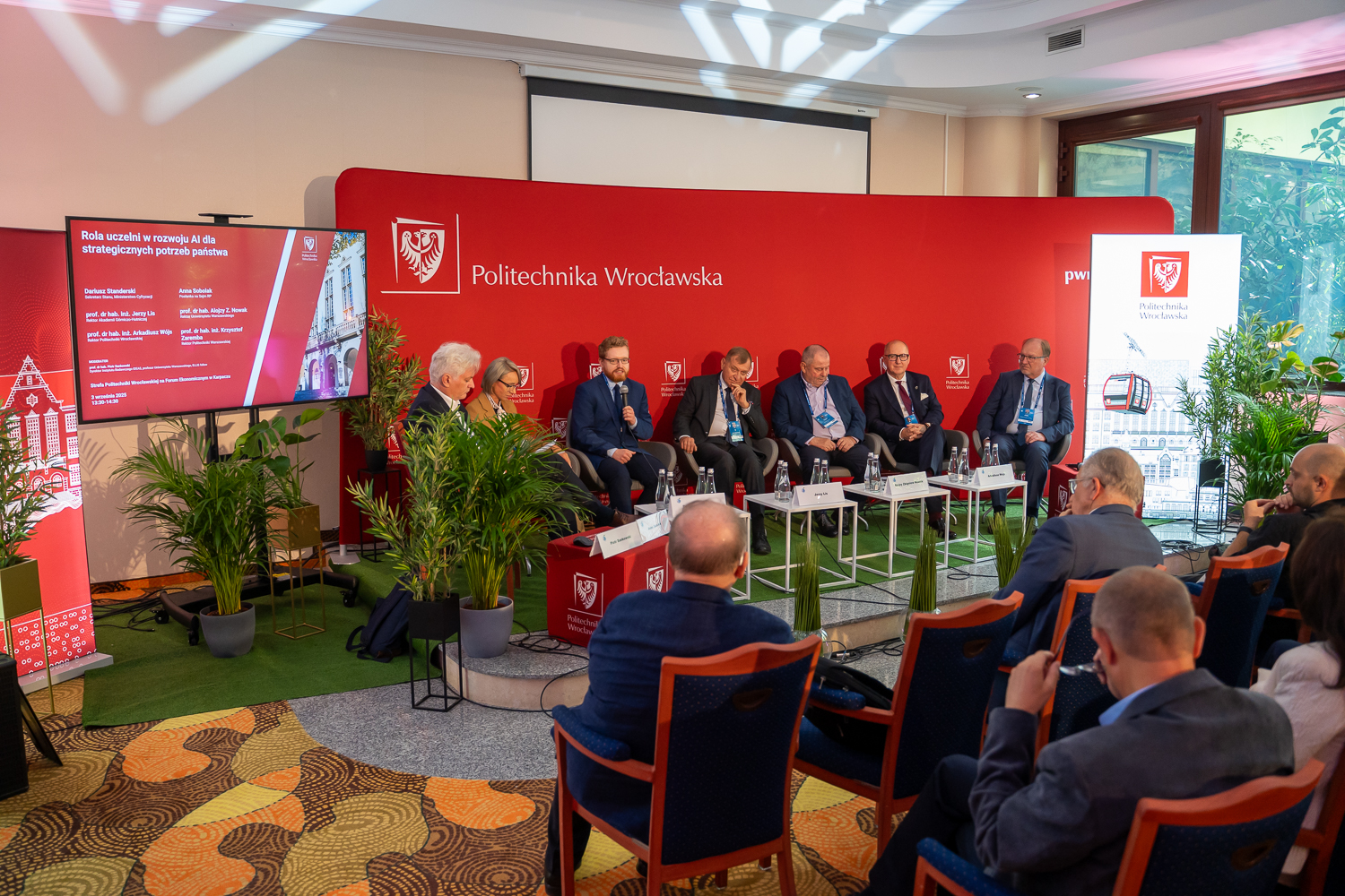 Uczestnicy panelu dyskusyjnego, w tym przedstawiciele Politechniki Wrocławskiej, siedzą na scenie przed czerwonym tłem z logo uczelni. Tłum słuchaczy obserwuje dyskusję, a na ekranie widoczny jest tytuł debaty "Rola uczelni w rozwoju AI dla strategicznych potrzeb państwa".