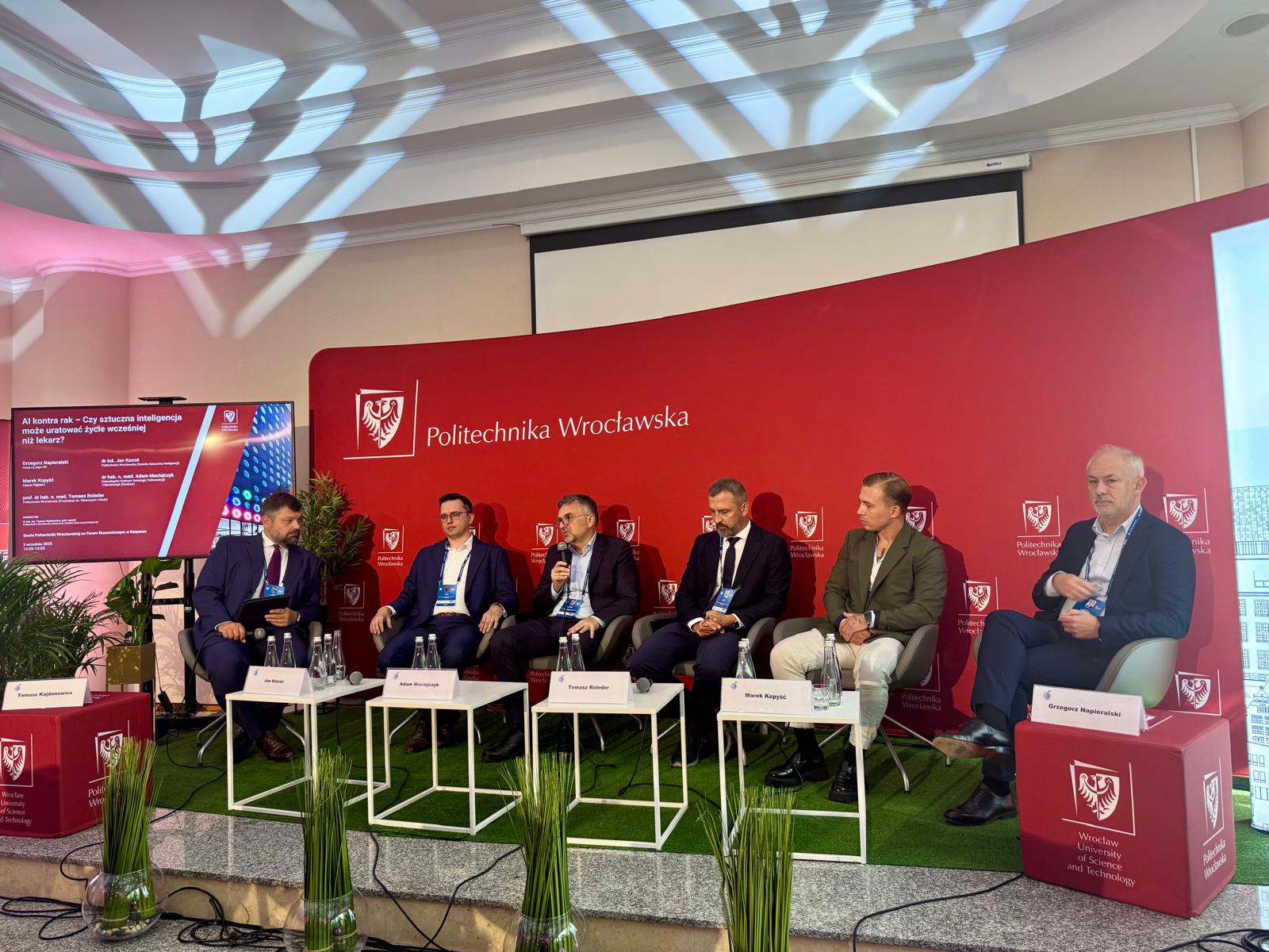 Sześciu panelistów, w tym mężczyźni w garniturach, siedzi na scenie w rzędzie, dyskutując przed czerwoną ścianką z logo Politechniki Wrocławskiej. Rośliny doniczkowe zdobią scenę. Na ekranie w tle widać tytuł debaty. Na stolikach leżą karteczki z nazwiskami.