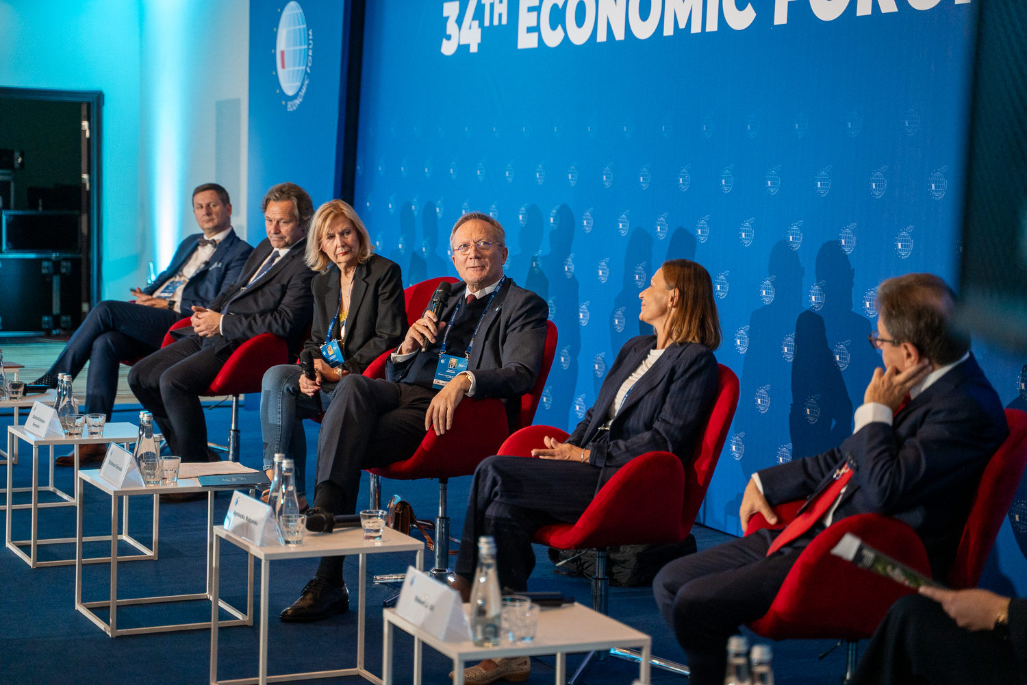 Grupa osób siedzi na czerwonych krzesłach panelowych, rozmawiając podczas 34. Forum Ekonomicznego. Mężczyzna w centrum trzyma mikrofon.