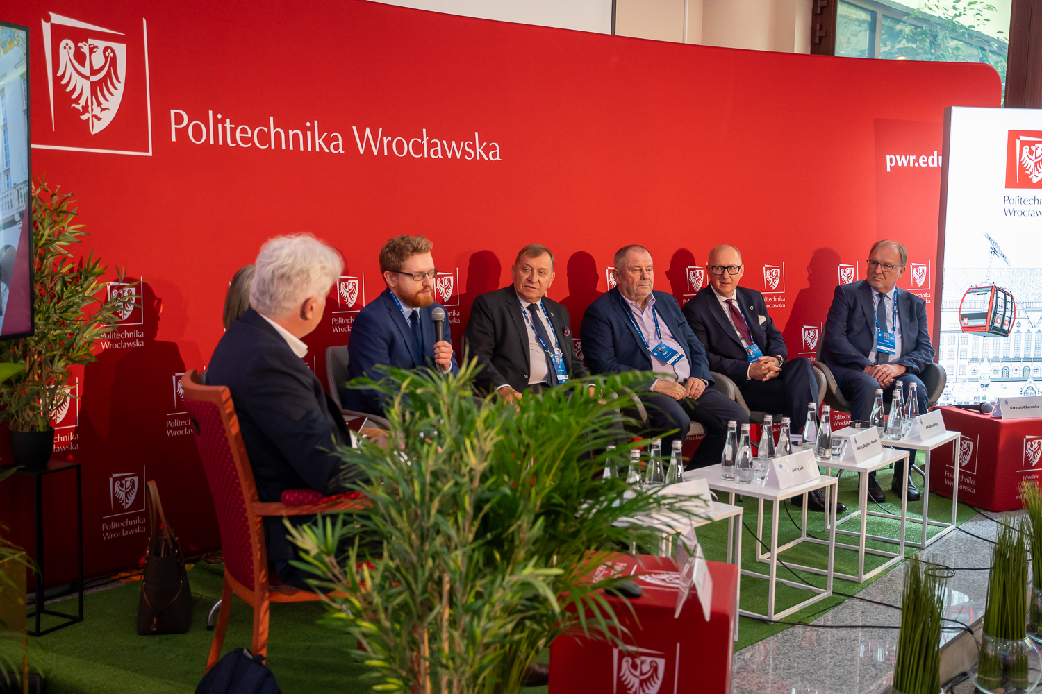 Grupa siedmiu panelistów, w tym kilku mężczyzn w garniturach, siedzi na scenie w rzędzie, z czerwoną ścianką w tle. Na banerach widoczne jest logo Politechniki Wrocławskiej. Rośliny doniczkowe dodają zieleni po bokach i na pierwszym planie.