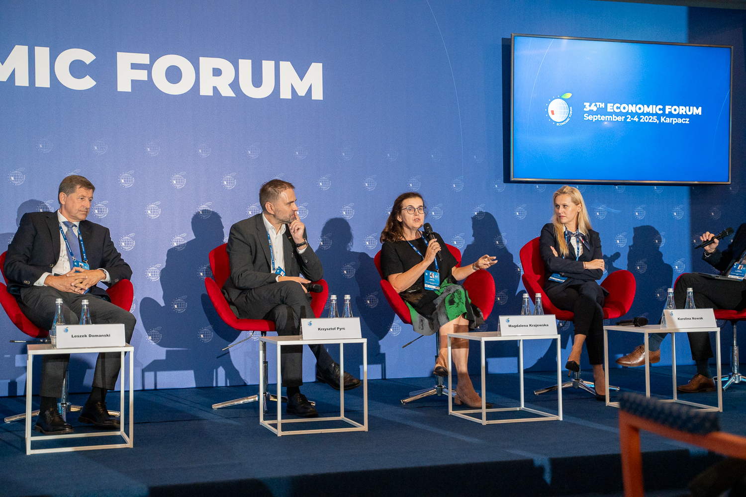 Czworo elegencko ubranych panelistów siedzi na tle niebieksiej ścianki. Jedna osoba trzyma mikrofon, a trzy pozostałem jej słuchają. Przed nimi stoją stoliki, na których są karteczki z nazwiskami.