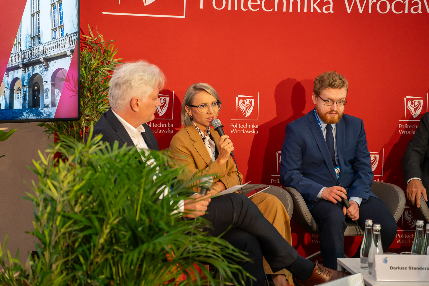 Panelistka w beżowym garniturze i okularach przemawia do mikrofonu, siedząc na scenie. Obok niej siedzą dwaj mężczyźni. W tle widoczne są czerwone banery z logo Politechniki Wrocławskiej.