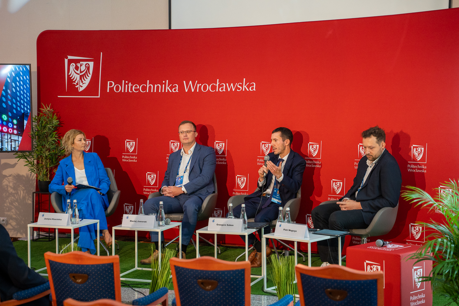 Czterech panelistów, w tym kobieta w niebieskim garniturze i trzech mężczyzn w garniturach, siedzi na scenie w rzędzie, z czerwoną ścianką w tle. Mężczyzna w środku trzyma mikrofon i mówi. W tle widać logo Politechniki Wrocławskiej. Na stolikach leżą karteczki z nazwiskami
