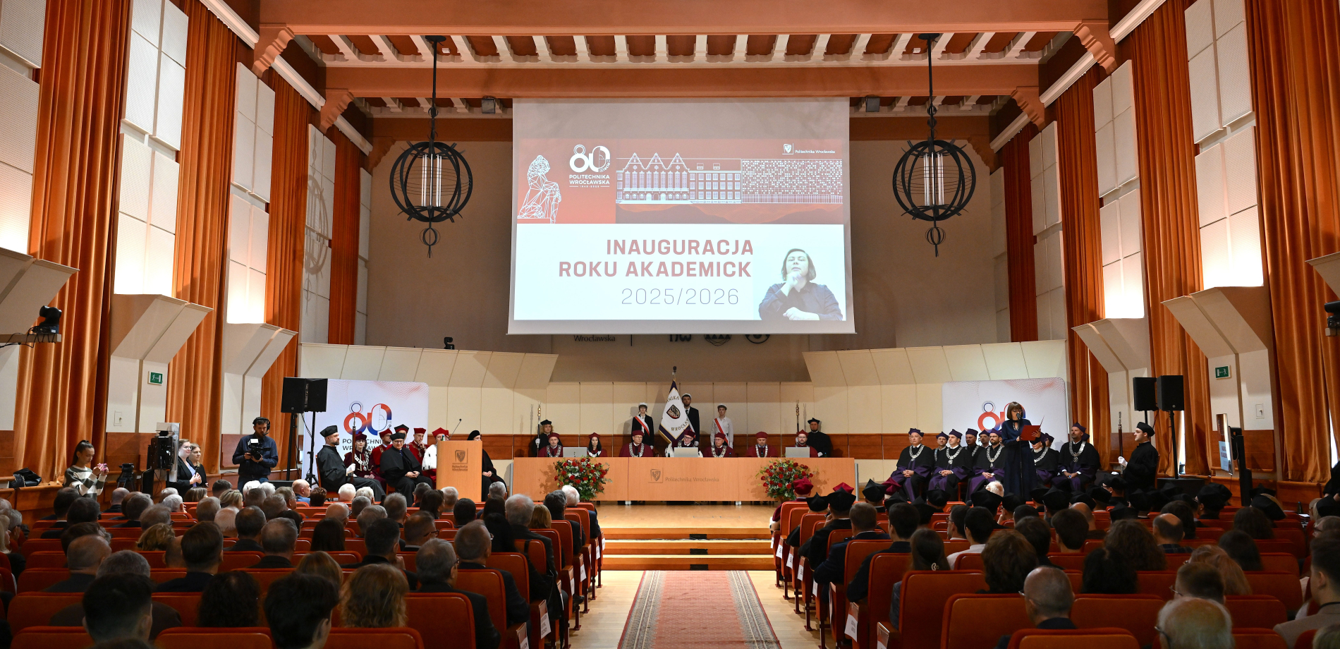 Duża sala wypełniona siedzącymi ludźmi. Na ekranie rzutnika widać napis Inauguracja roku akademickiego 2025/26.