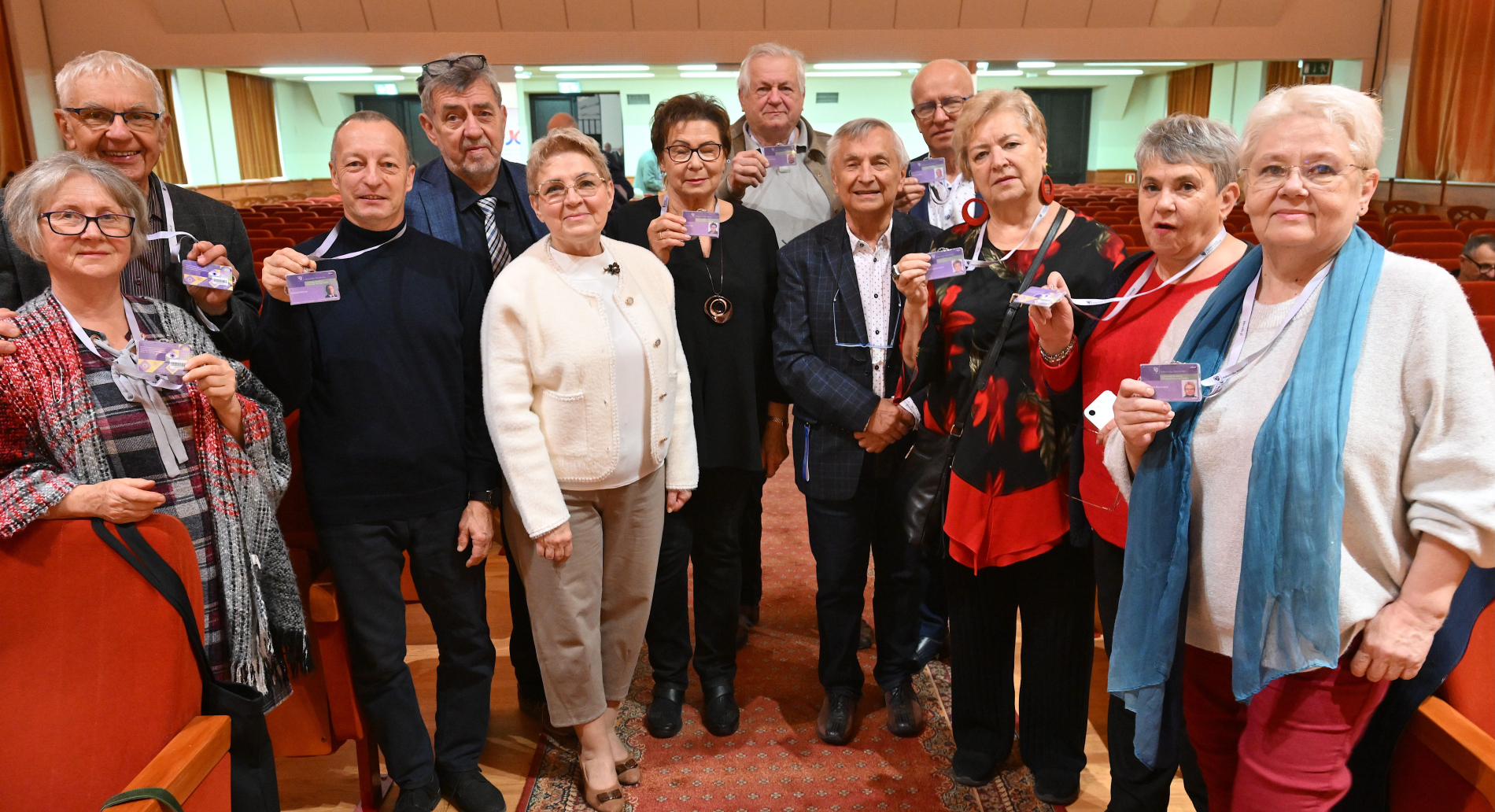 Grupa kilkunastu uśmiechniętych, starszych osób pozuje w sali wykładowej. Wszyscy trzymają w rękach identyfikatory studenckie.