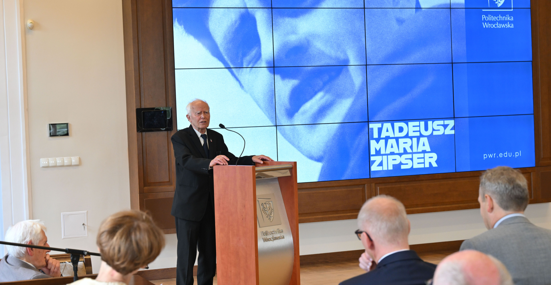 Starszy mężczyzna w garniturze przemawia z mównicy, w tle duży niebieski ekran z portretem i nazwiskiem Tadeusza Marii Zipsera oraz logo Politechniki Wrocławskiej.