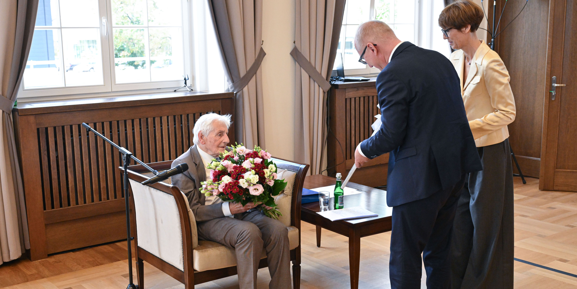 Starszy mężczyzna siedzi na fotelu z dużym bukietem kwiatów, uśmiecha się. Przed nim stoi mężczyzna w garniturze i kobieta.