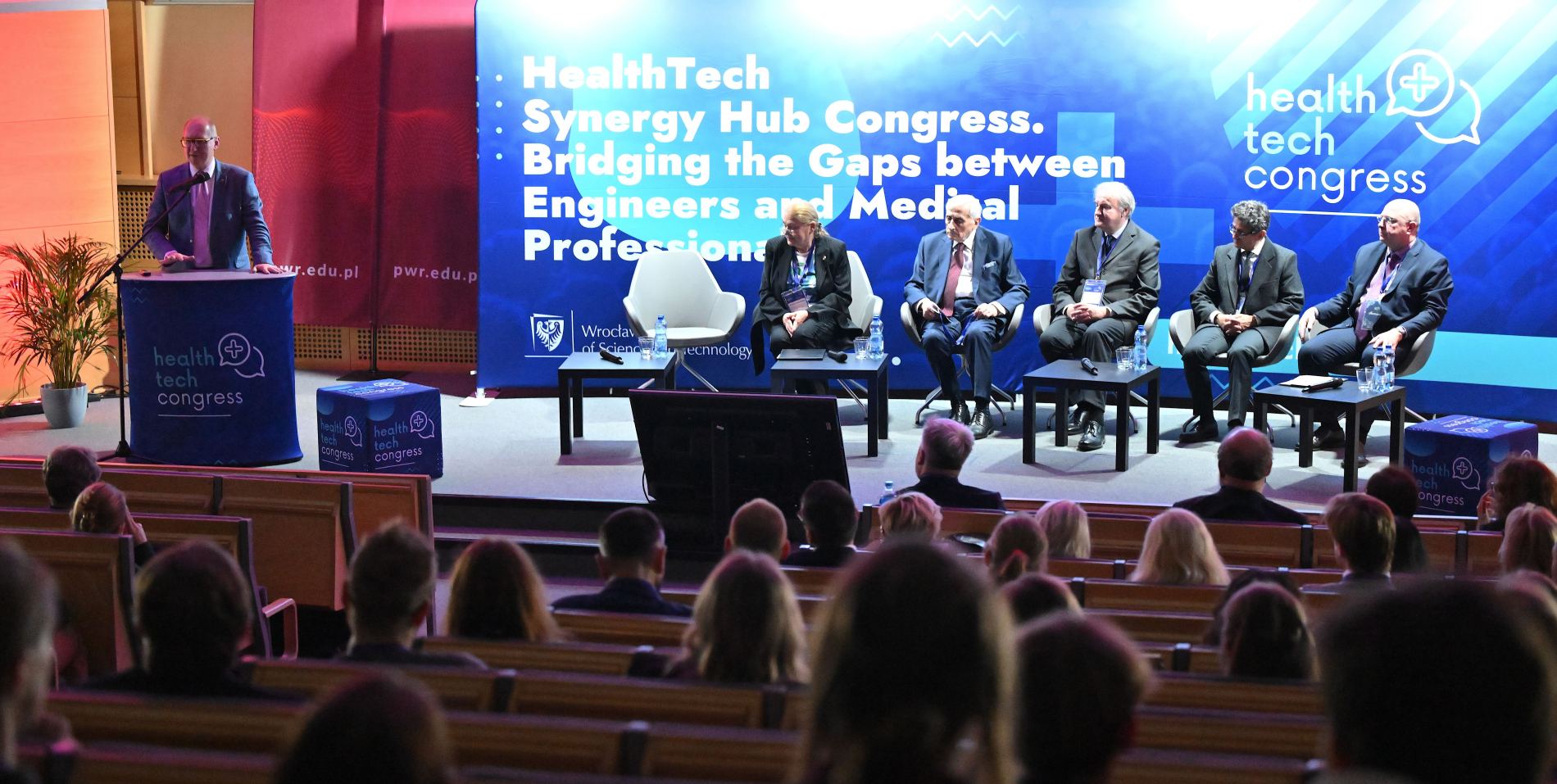 Prelegent przemawia z podium na scenie podczas "Health Tech Congress". Na scenie siedzi pięciu panelistów. W tle duży ekran z tytułem wydarzenia.