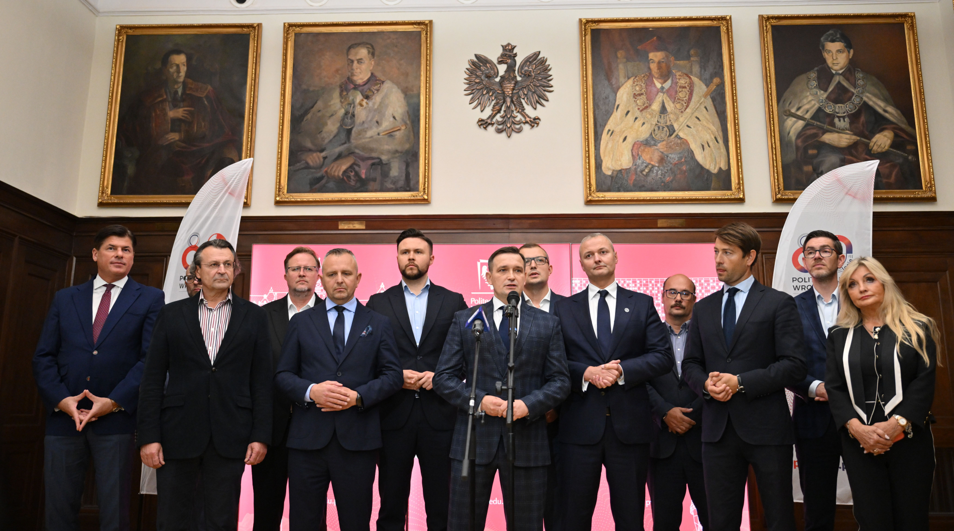 Na zdjęciu widoczna jest grupa oficjalnie ubranych osób, stojąca w rzędzie. W tle znajdują się portrety w złotych ramach oraz godło Polski, a po bokach białe banery.