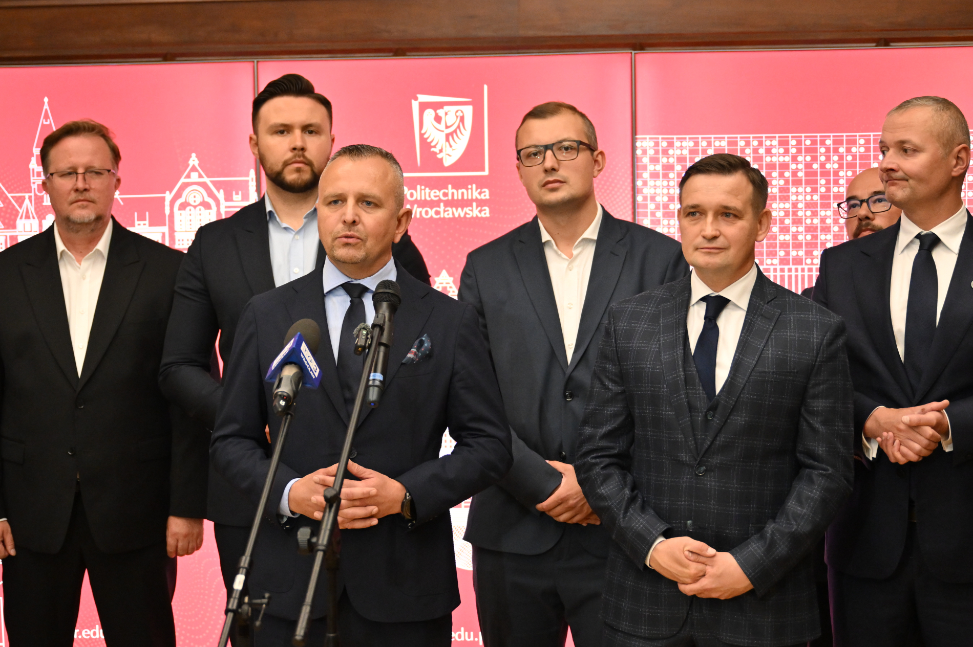 Grupa mężczyzn w garniturach stoi przed czerwoną ścianką z logo Politechniki Wrocławskiej. Centralnie ustawiony mężczyzna z krótkimi siwiejącymi włosami przemawia do mikrofonów. Pozostali stoją obok niego, patrząc w kierunku kamery.