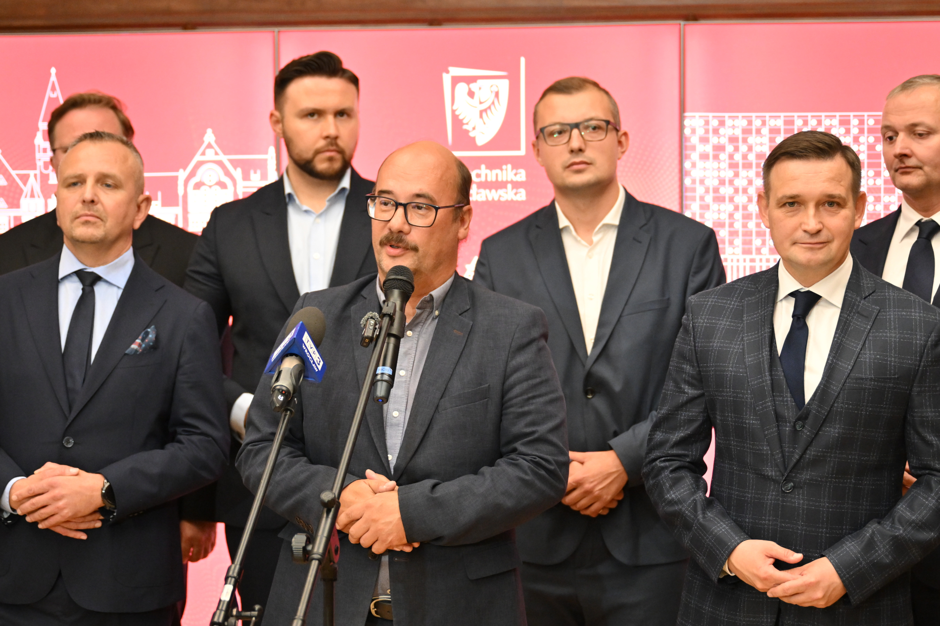 Grupa mężczyzn, ubranych w garnitury, stoi przed czerwoną ścianką z logo Politechniki Wrocławskiej. W centrum kadru mężczyzna z brodą i okularami przemawia do mikrofonów. Pozostali stoją obok, skierowani w stronę kamery.