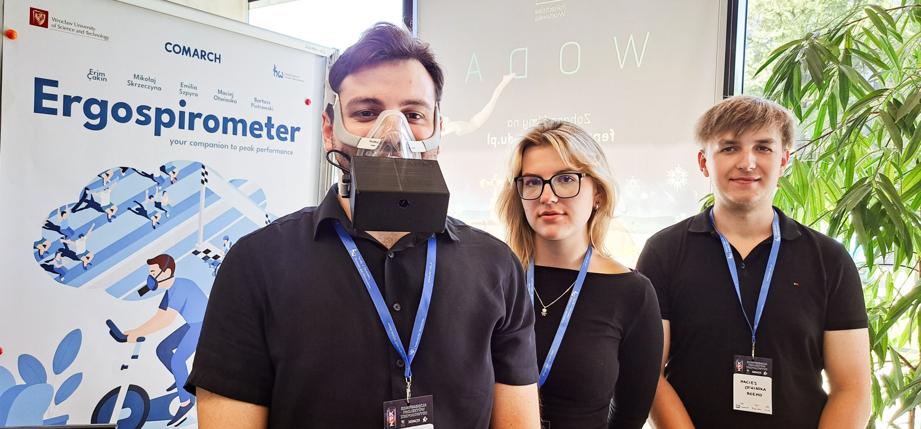 Trzy osoby stoją obok plakatu reklamującego "Ergospirometer Comarch". Mężczyzna po lewej stronie ma na sobie urządzenie przypominające maskę oddechową. Obok niego stoi kobieta w okularach, a po prawej kolejny mężczyzna. Wszyscy mają na sobie ciemne koszulki i smycze z identyfikatorami.
