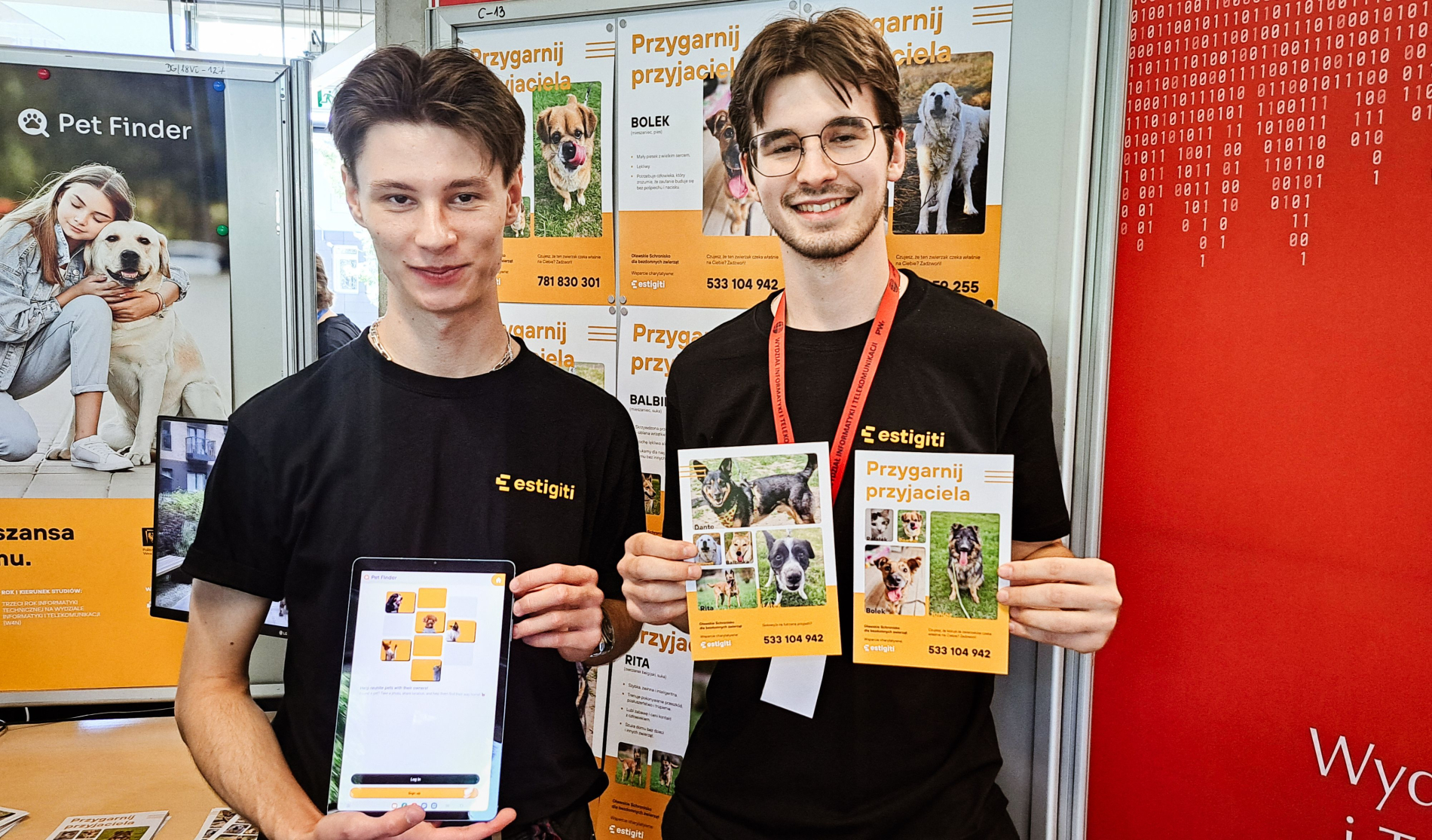 Dwóch młodych mężczyzn stoi przy stoisku "Pet Finder", prezentując materiały promocyjne. Jeden trzyma tablet z otwartą aplikacją, drugi ulotkę zatytułowaną "Przygarnij przyjaciela", przedstawiającą zdjęcia psów. W tle widać plakaty z podobną tematyką.