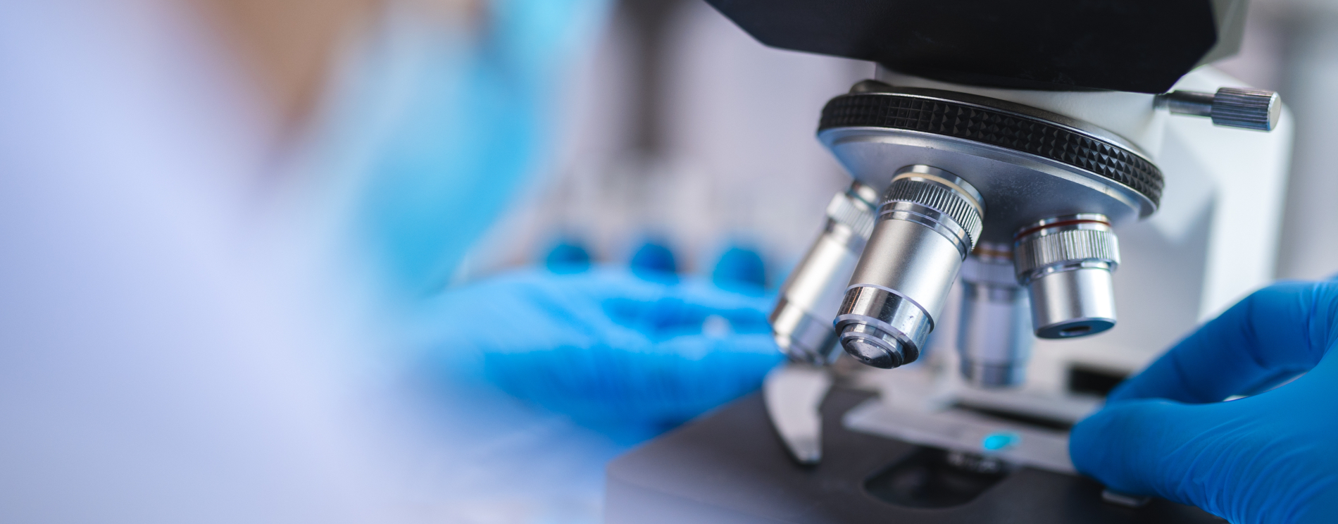 Zbliżenie na mikroskop laboratoryjny, przy którym naukowiec w niebieskich rękawiczkach umieszcza preparat na stoliku. W tle, niewyraźne postacie w rękawiczkach pracują w laboratorium.