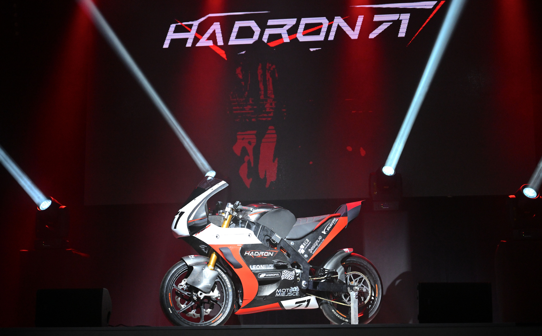 Motocykl wyścigowy Hadron w barwach biało-czerwono-czarnych na scenie podczas prezentacji. Nad nim duży, podświetlony na czerwono napis z nazwą modelu.