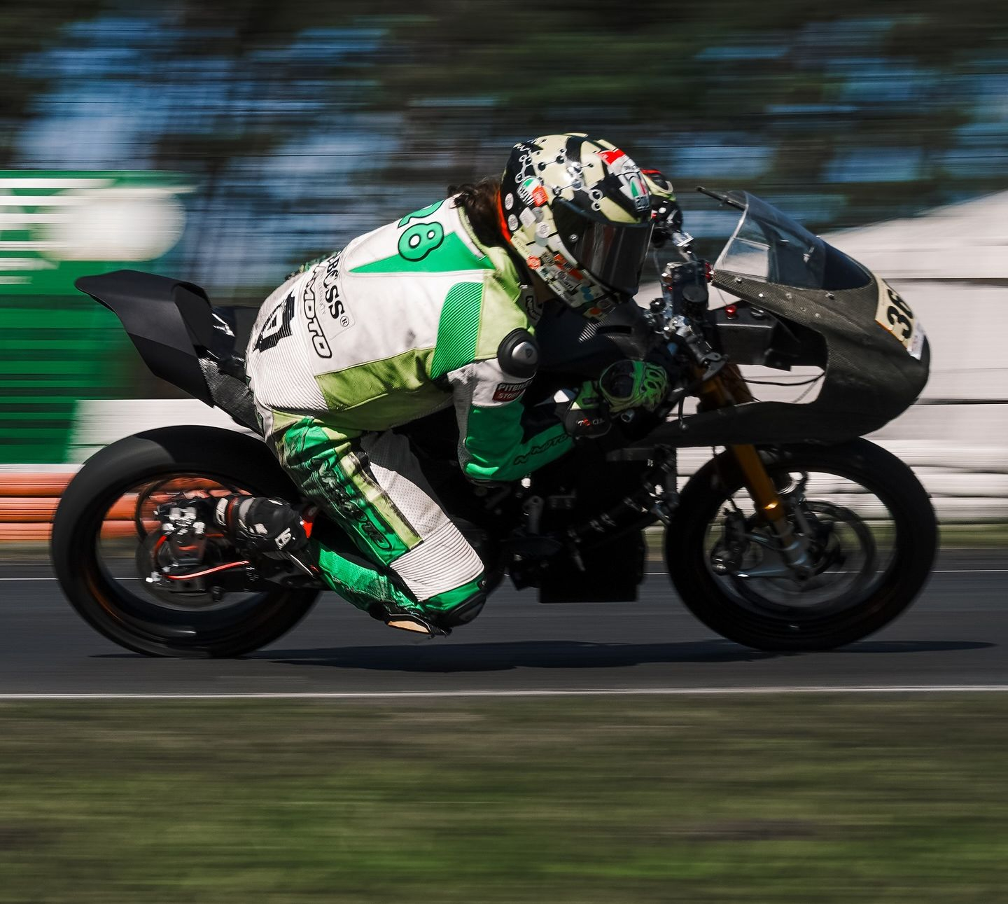 Ujęcie przedstawiające motocyklistę w zielono-białym kombinezonie i kasku, jadącego motocyklem sportowym na torze wyścigowym przy dużej prędkości.