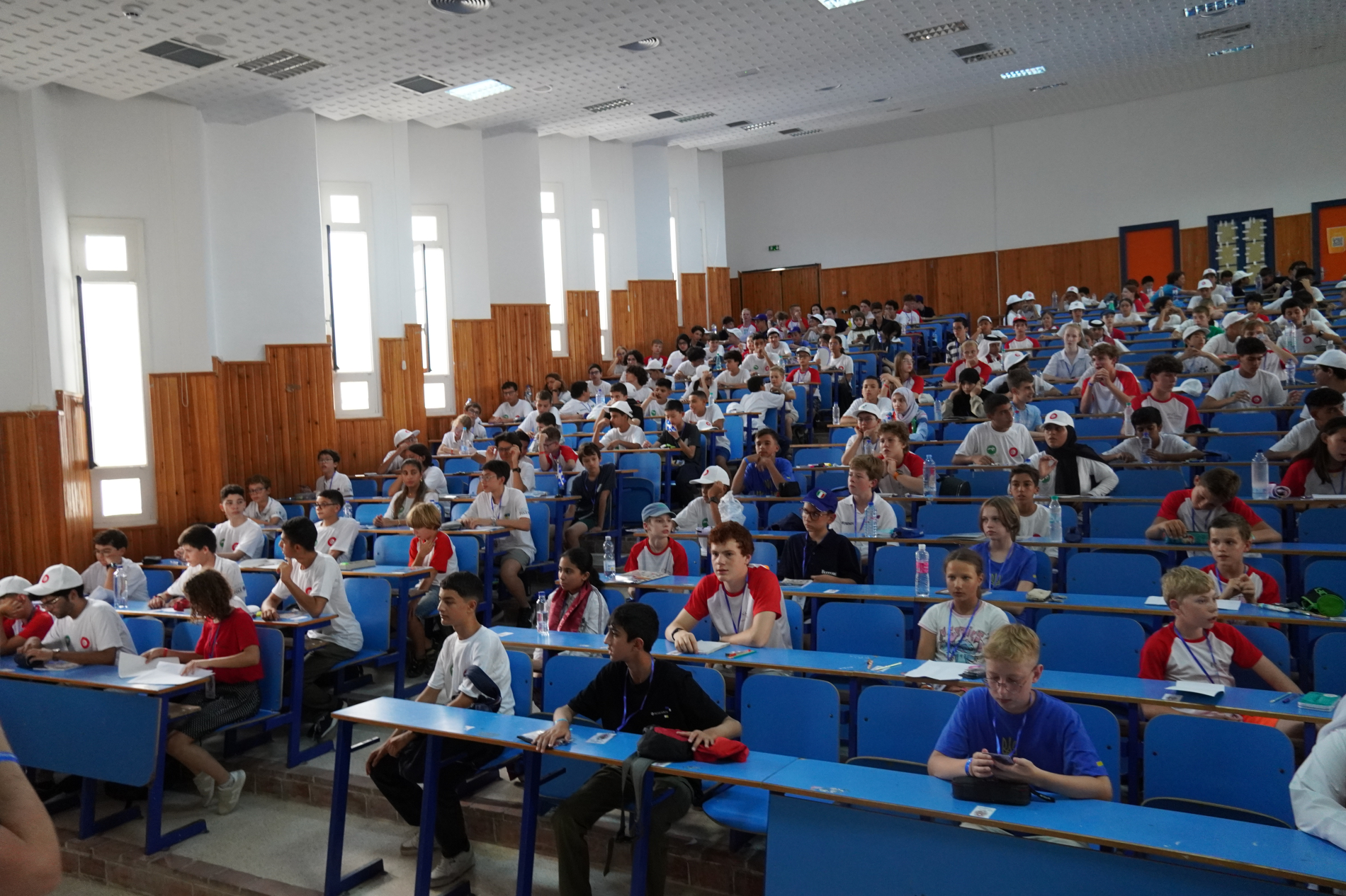 Zdjęcie zrobione w szerokiej auli, przedstawia dużą grupę młodych ludzi, prawdopodobnie uczniów, siedzących na niebieskich krzesłach przy niebieskich ławkach. Większość z nich ma na sobie białe koszulki z czerwonymi elementami. Z przodu widać uczniów zajętych notowaniem, a w głębi sali widać więcej osób siedzących w rzędach. Wnętrze jest dobrze oświetlone, ze ścianami w drewnie i białym tynkiem.