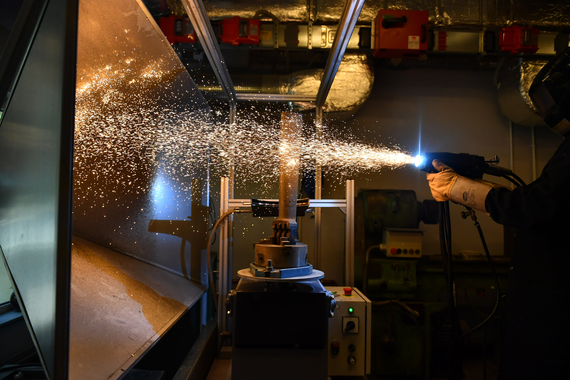 Proces natryskiwania cieplnego: pracownik w rękawicy kieruje palnik generujący snop iskier i światła na obracający się element maszynowy.