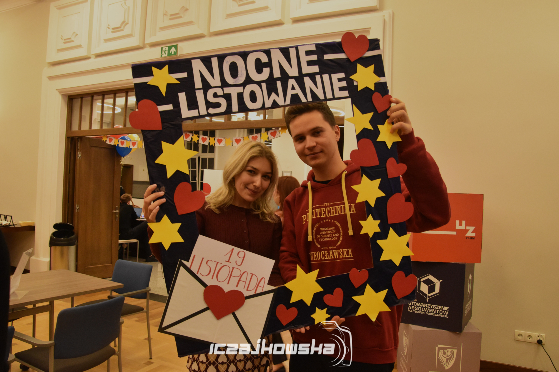 Młoda kobieta i młody mężczyzna trzymają dużą, prostokątną ramkę ozdobioną sercami i gwiazdami z napisem "Nocne Listowanie".