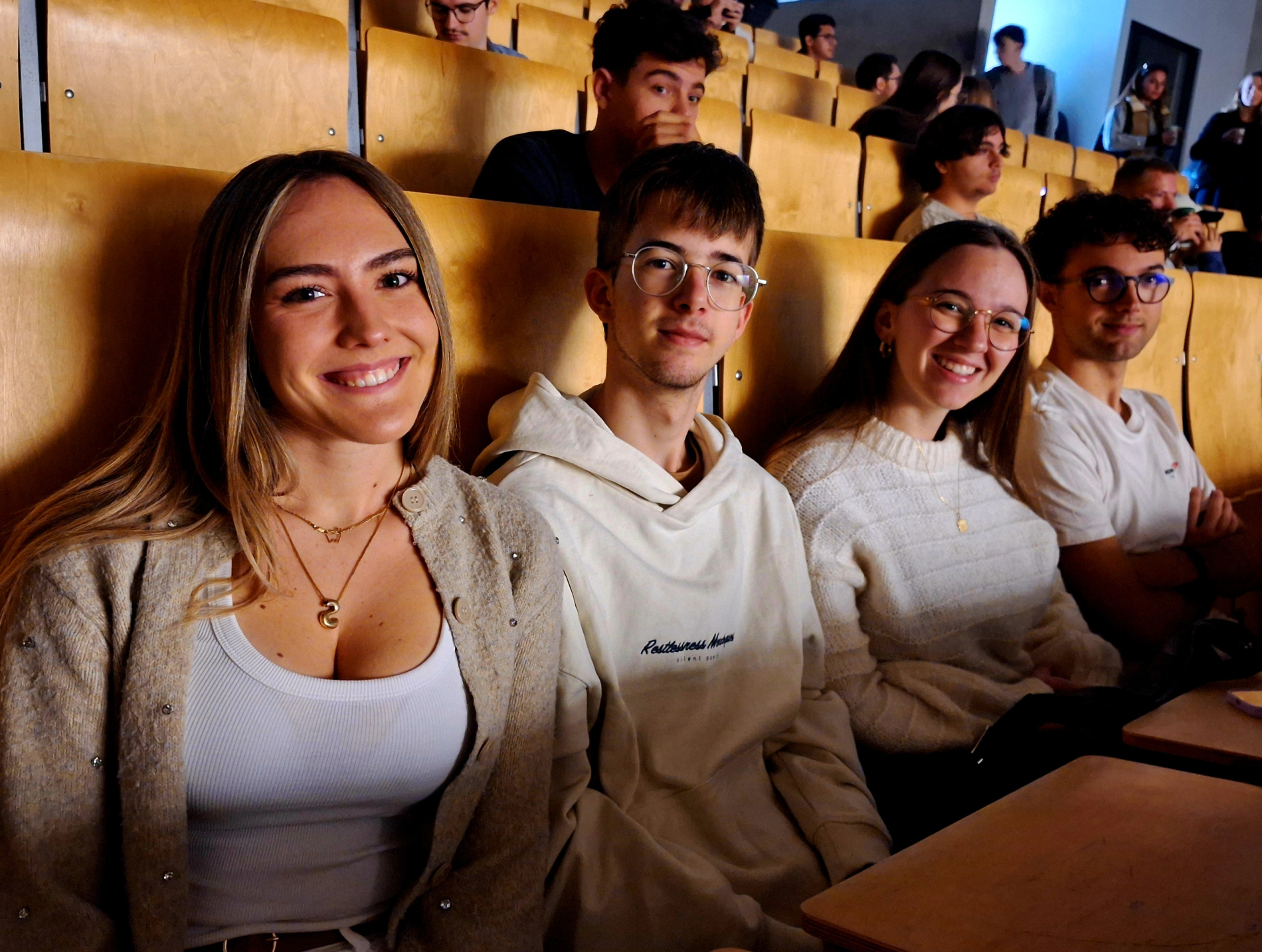 Czworo uśmiechniętych, młodych studentów siedzących razem w pierwszym rzędzie w drewnianej auli. 