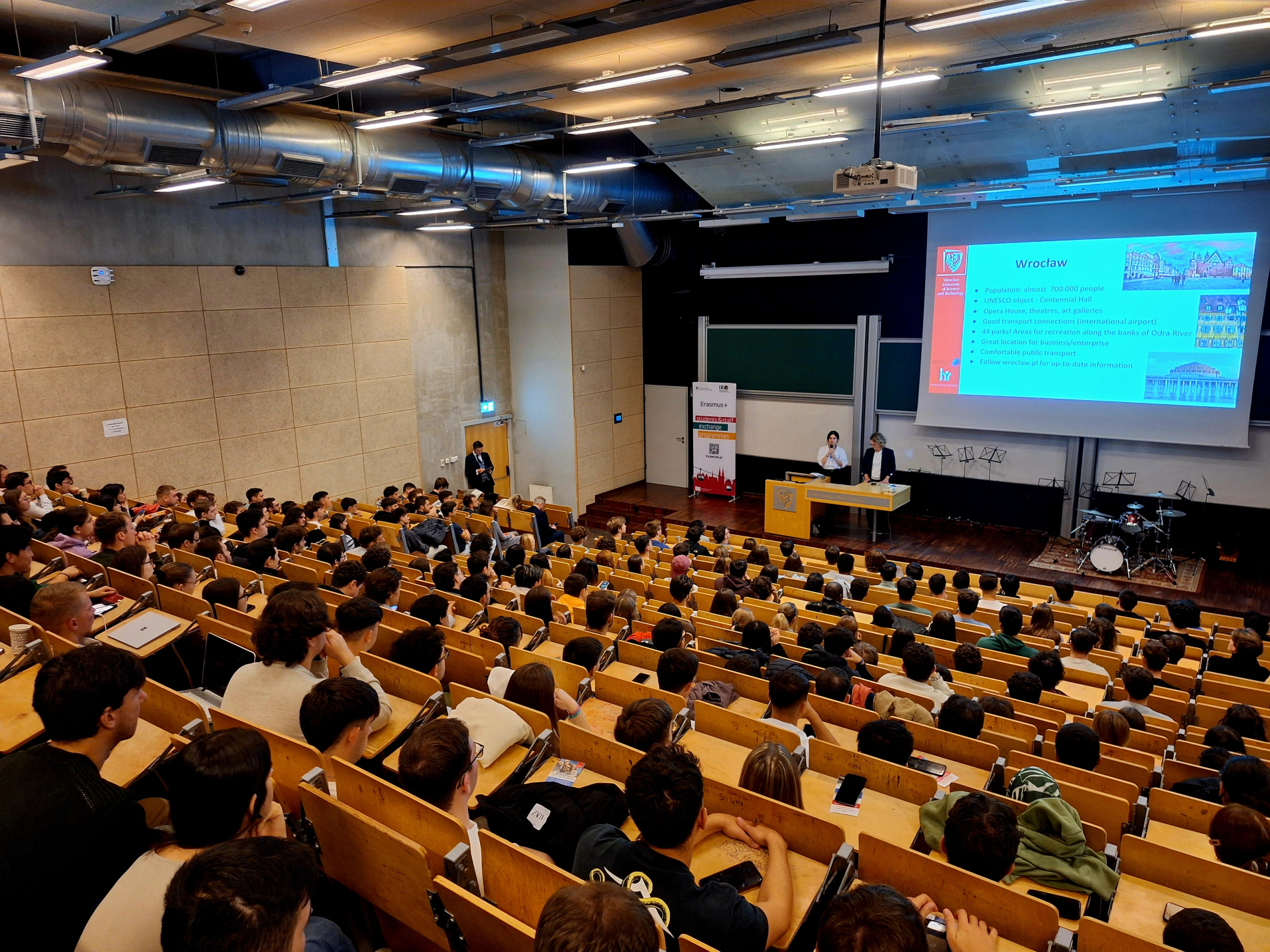 Szeroki widok na dużą, nowoczesną aulę wypełnioną studentami, słuchającymi wykładu; na ekranie slajd z napisem "Wrocław".