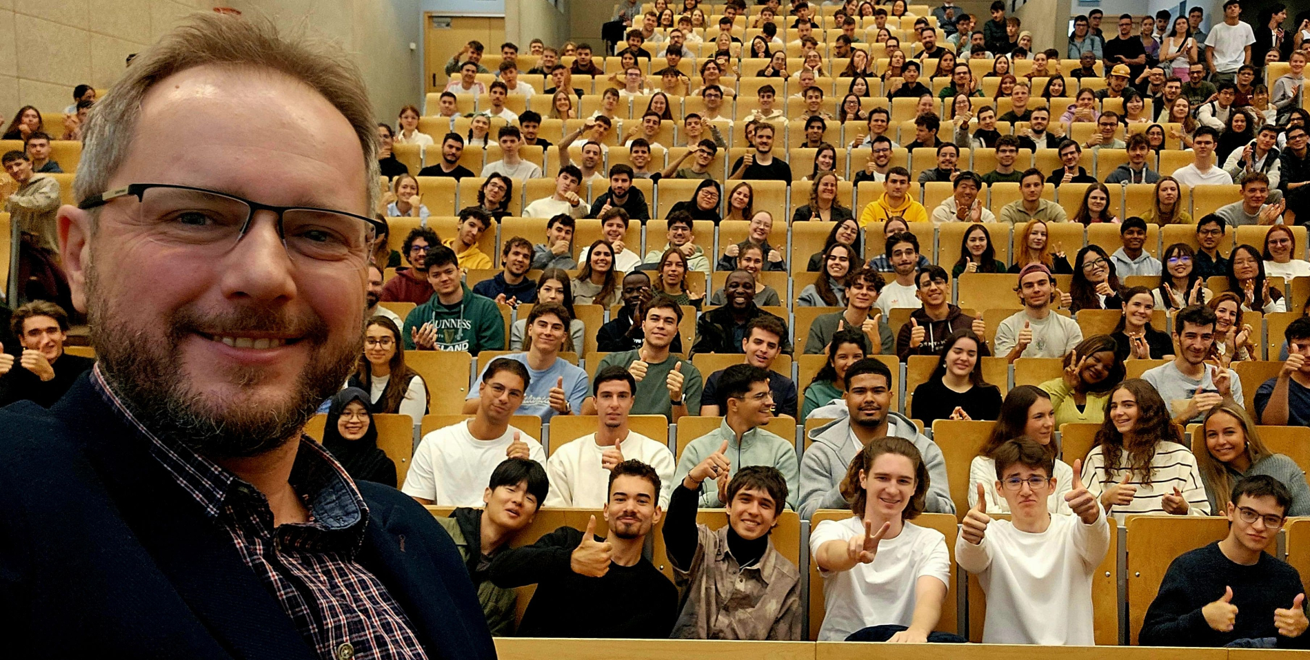 Uśmiechnięty, brodaty mężczyzna w okularach (selfie z bliska) na tle wypełnionej studentami auli, którzy pokazują kciuki w górę.