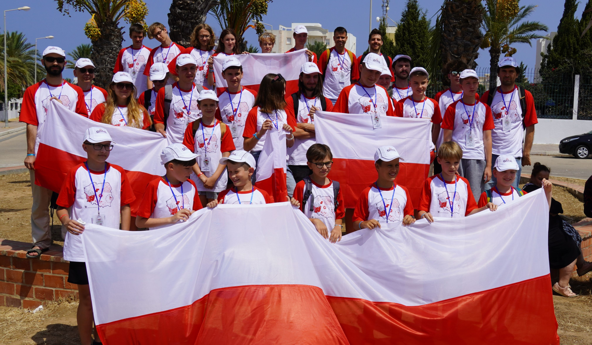 Zdjęcie przedstawia dużą grupę młodych ludzi i kilku dorosłych, uśmiechniętych i trzymających biało-czerwoną flagę Polski. Wszyscy ubrani są w białe koszulki z czerwonymi rękawami, ozdobione grafiką, oraz białe czapki z daszkiem. Stoją na zewnątrz, w słoneczny dzień, w tle widać palmy i budynki.