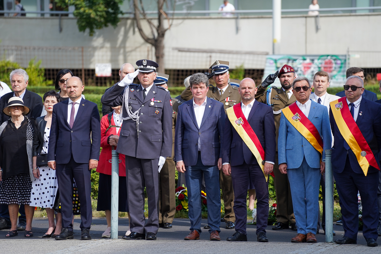 Grupa mężczyzn w mundurach i eleganckich garniturach, z szarfami orderowymi na piersiach, stoi na placu podczas uroczystości. W tle widać drzewa i zarys budynku.