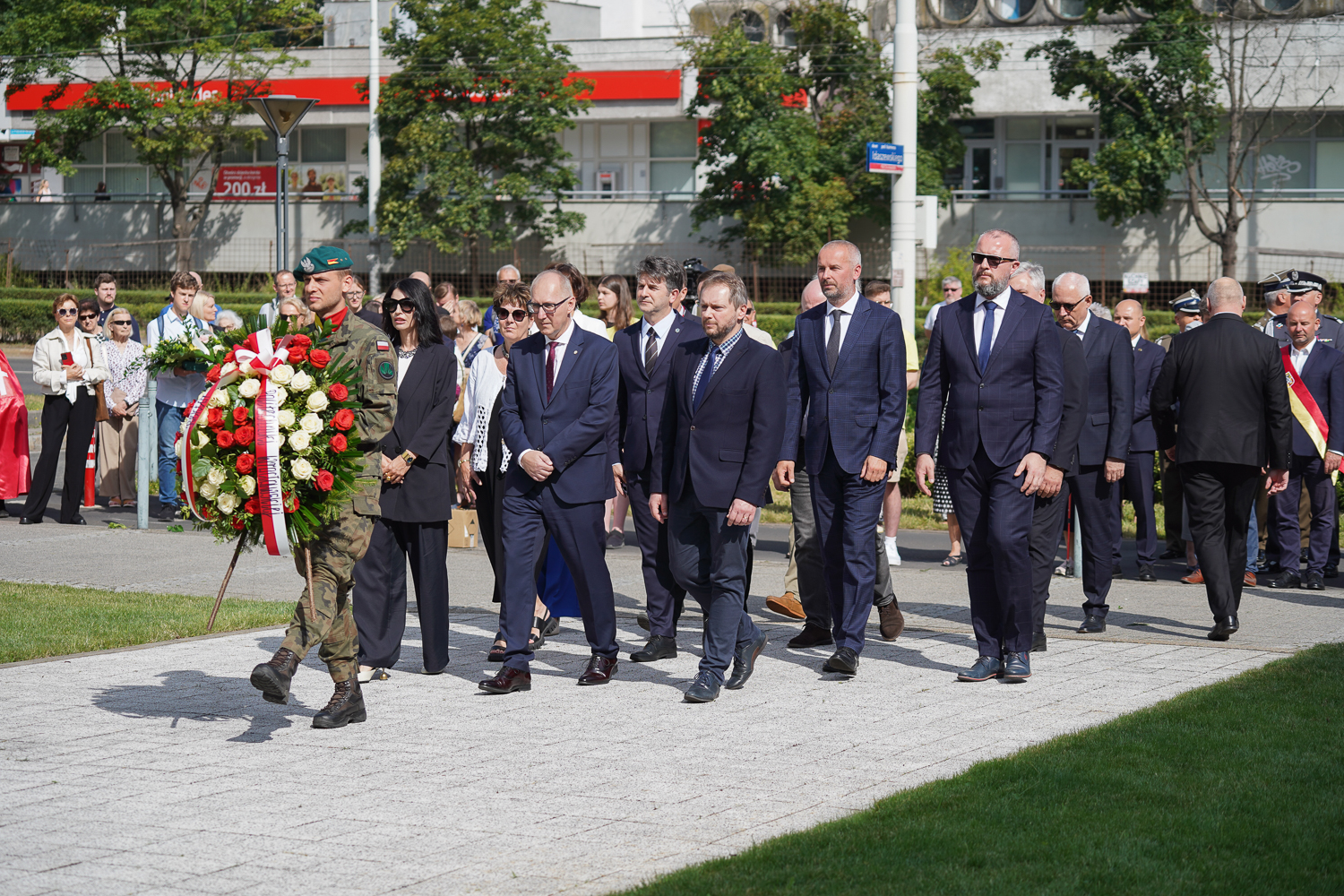 Żołnierz w mundurze z wieńcem biało-czerwonych kwiatów, idący na czele grupy osób w formalnych strojach. W tle budynki miejskie i drzewa, sugerujące ulicę.
