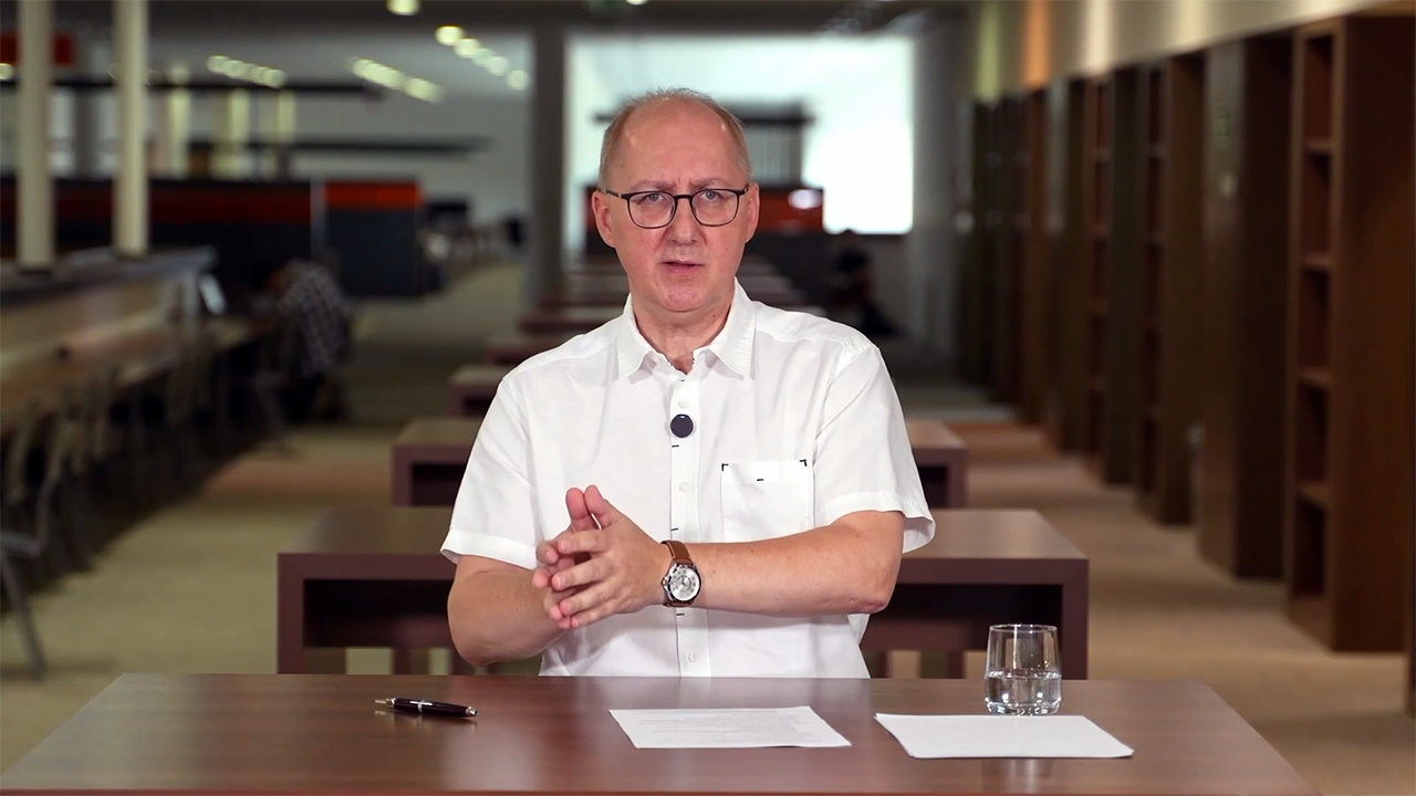 Mężczyzna w okularach i białej koszuli siedzi przy stole, gestykulując. Przed nim leżą kartki i długopis oraz szklanka wody. W tle widać regały z książkami.