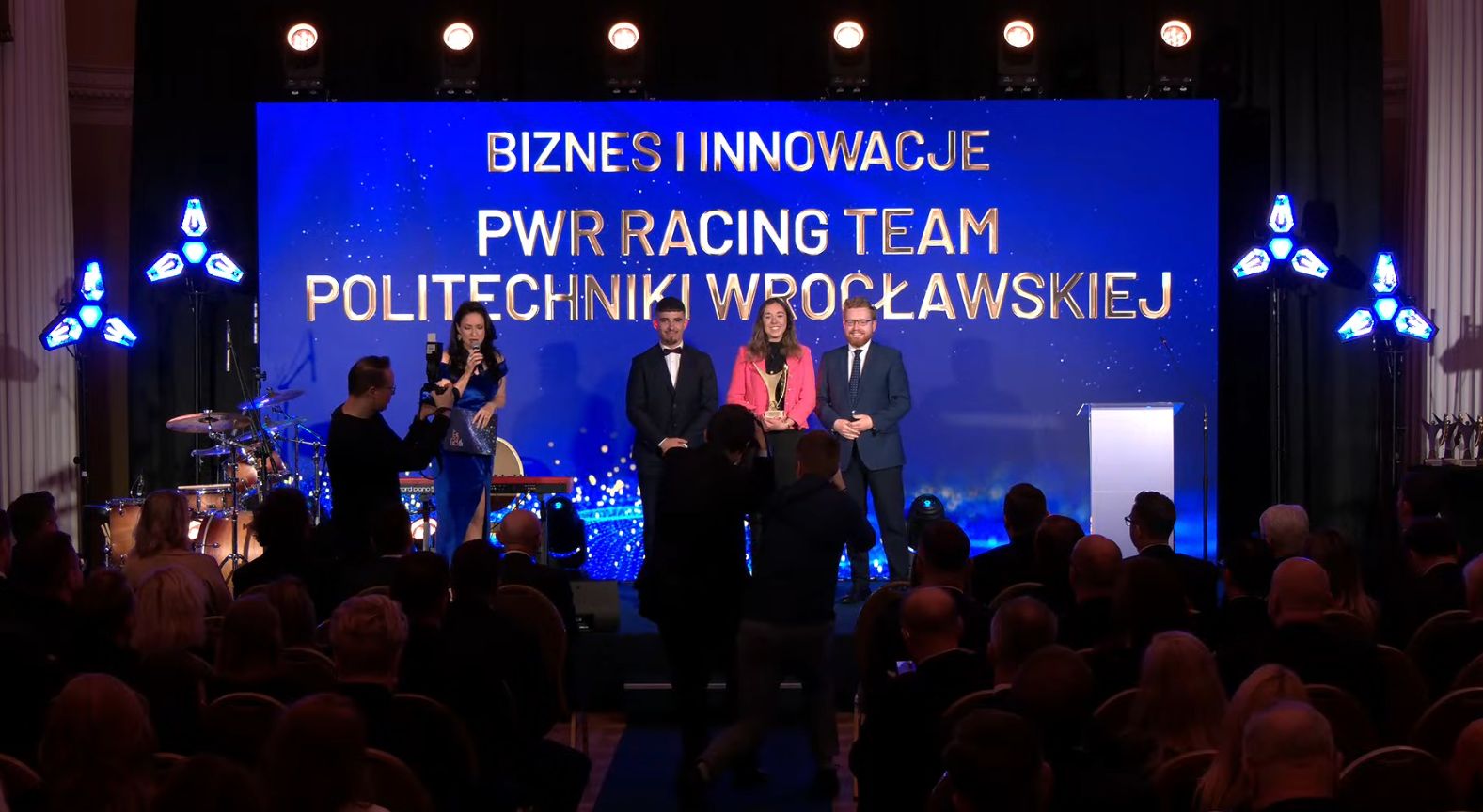 Uroczystość wręczenia nagród: na scenie 4 osoby (dwie kobiety, dwóch mężczyzn) przed dużym ekranem.