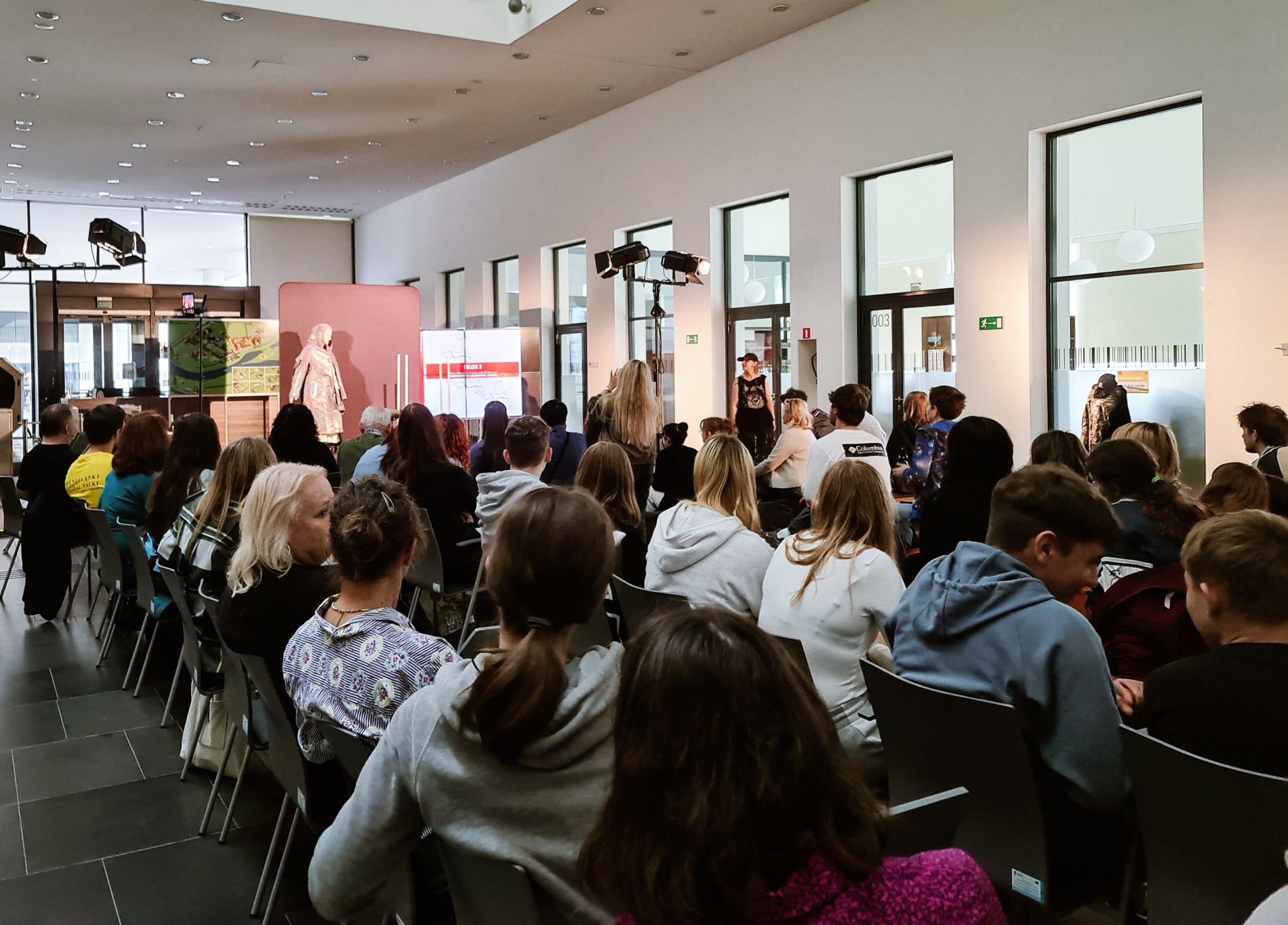 Grupa ludzi siedzi na krzesłach w rzędach, słuchając prelegentki. Kobieta w jasnym kapeluszu i jasnej bluzce stoi przed publicznością, prowadząc prezentację. W tle widoczny jest plakat oraz duża rzeźba.