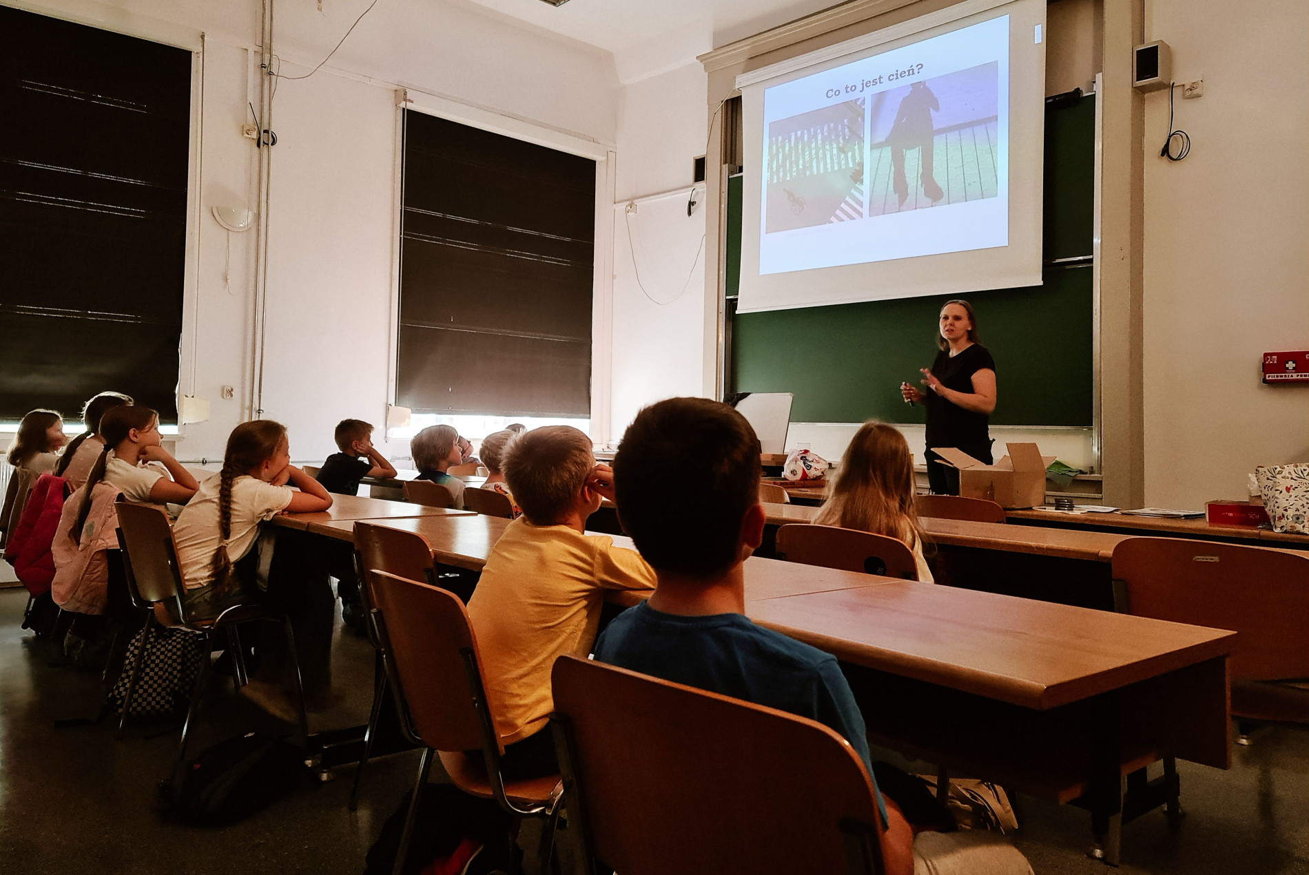 Nauczycielka w ciemnym ubraniu stoi przed tablicą, prowadząc zajęcia dla grupy dzieci. Na ekranie projektora widoczny jest slajd z pytaniem "Co to jest cień?". Dzieci siedzą w ławkach, słuchając wykładu.