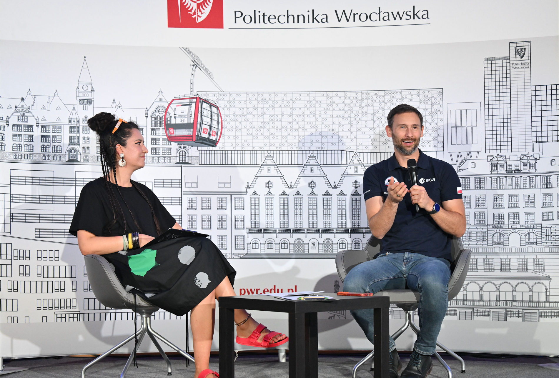 Sławosz Uznański w granatowej koszulce i kobieta w czarnej sukience z białymi kropkami siedzą na krzesłach, prowadząc rozmowę. Sławosz trzyma mikrofon i gestykuluje. W tle rysunek Wrocławia.