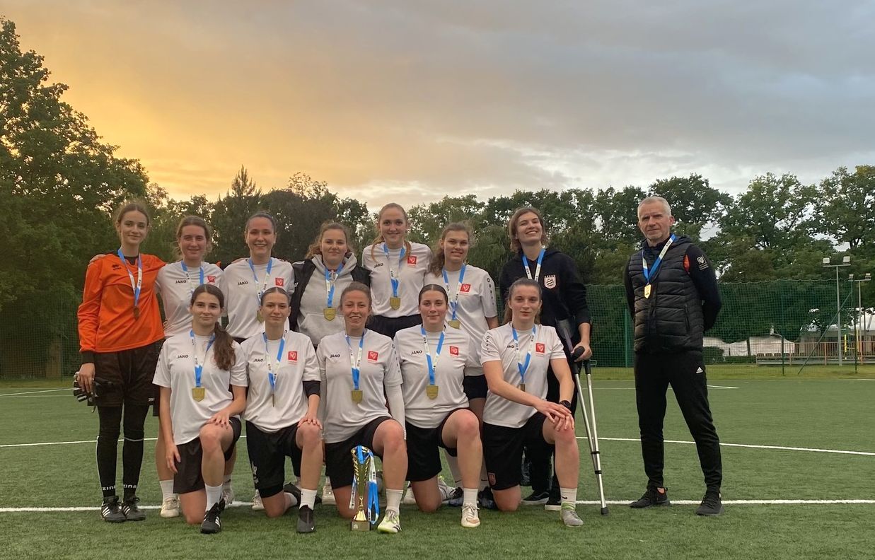 Zdjęcie grupowe drużyny piłkarskiej kobiet z medalami i pucharem, stojących na boisku. W tle widać drzewa i zachodzące słońce. Obok zawodniczek stoi trener.