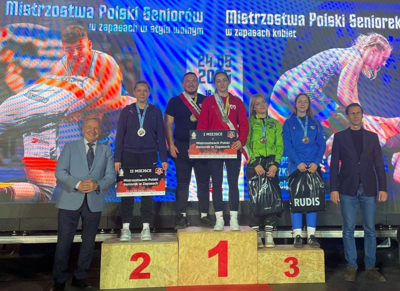 Zdjęcie przedstawia podium Mistrzostw Polski Seniorów w zapasach. Na najwyższym stopniu stoją zwycięzcy, obok nich osoby na 2. i 3. miejscu. W tle widoczne są grafiki przedstawiające zapaśników.